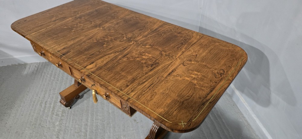 A Regency rosewood sofa table with two frieze drawers, brass line inlay, drop leaves, carved rosette corners, and reeded legs on brass castors. View X