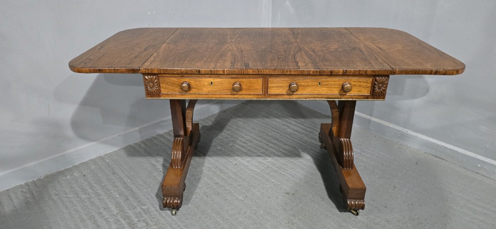 A Regency rosewood sofa table with two frieze drawers, brass line inlay, drop leaves, carved rosette corners, and reeded legs on brass castors. View XXI
