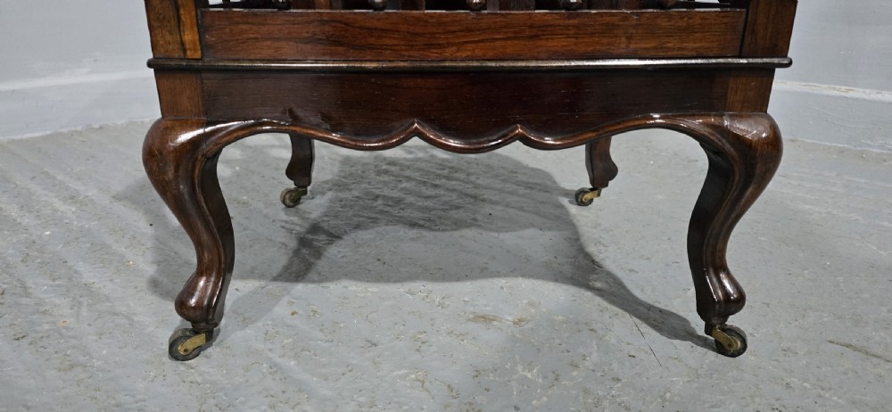 A dark rosewood Victorian Canterbury featuring turned spindle sides, vertical dividers, a flat top surface, and cabriole legs on brass castors. View VI