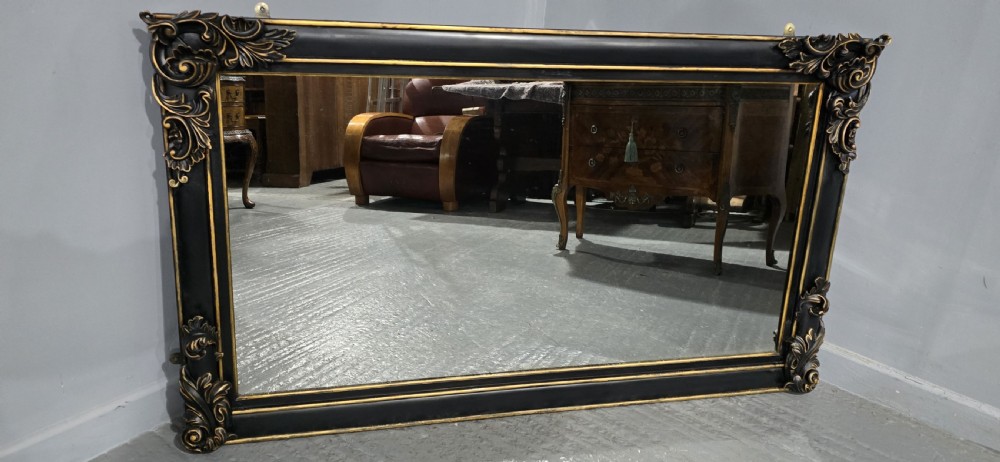 A large horizontal rectangular overmantel mirror with an ebonised black wooden frame, accented by gilt inner moldings and detailed foliate corner flourishes. View II