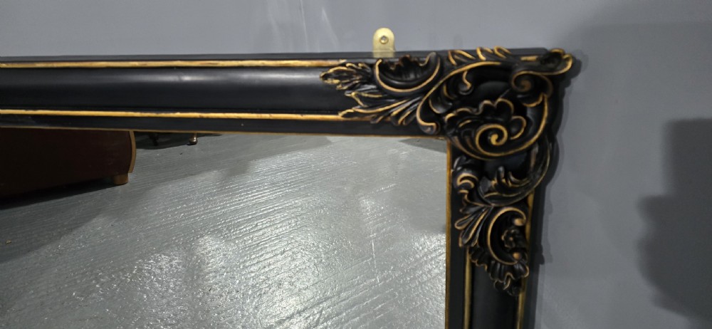 A large horizontal rectangular overmantel mirror with an ebonised black wooden frame, accented by gilt inner moldings and detailed foliate corner flourishes. View VII