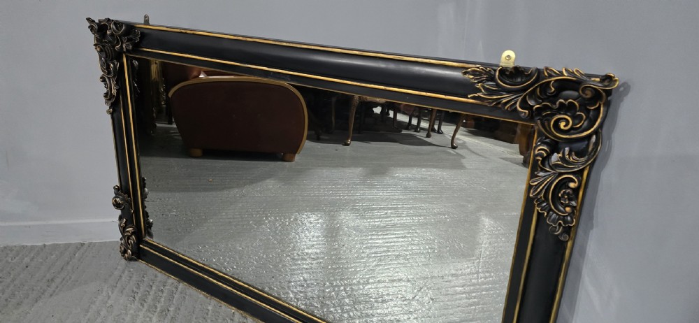 A large horizontal rectangular overmantel mirror with an ebonised black wooden frame, accented by gilt inner moldings and detailed foliate corner flourishes. View IX