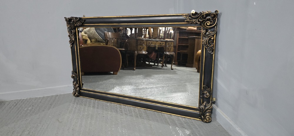 A large horizontal rectangular overmantel mirror with an ebonised black wooden frame, accented by gilt inner moldings and detailed foliate corner flourishes. View X