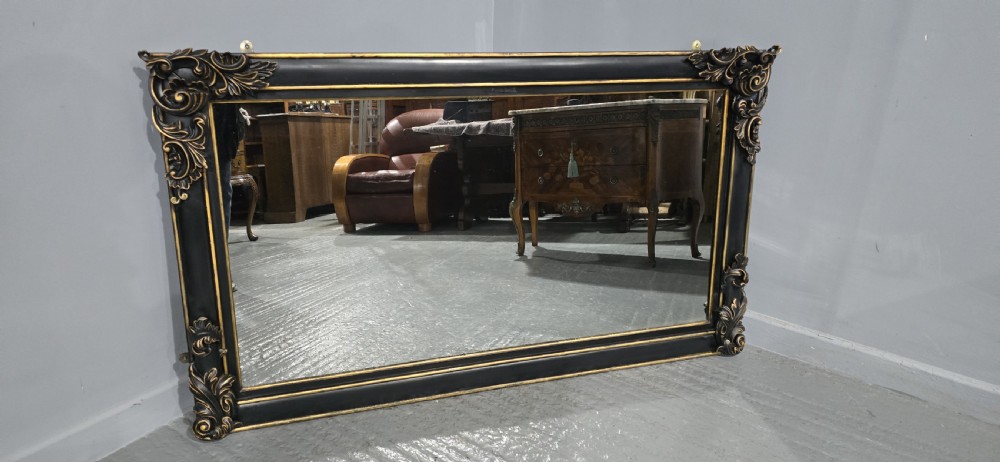 A large horizontal rectangular overmantel mirror with an ebonised black wooden frame, accented by gilt inner moldings and detailed foliate corner flourishes. View XI