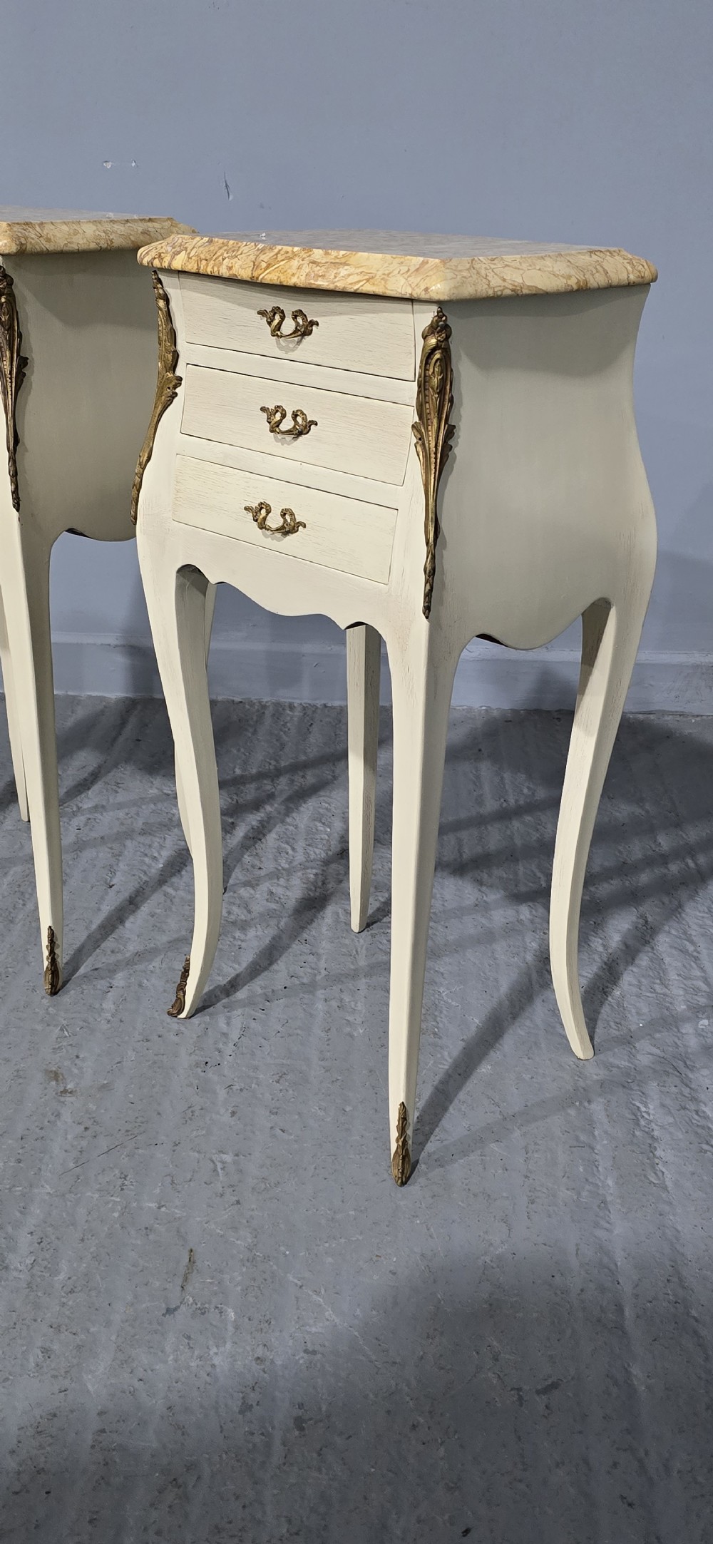 A pair of slender cream-painted French Louis style bedside chests featuring shaped marble tops, three drawers, and decorative brass mounts on tall cabriole legs. View III