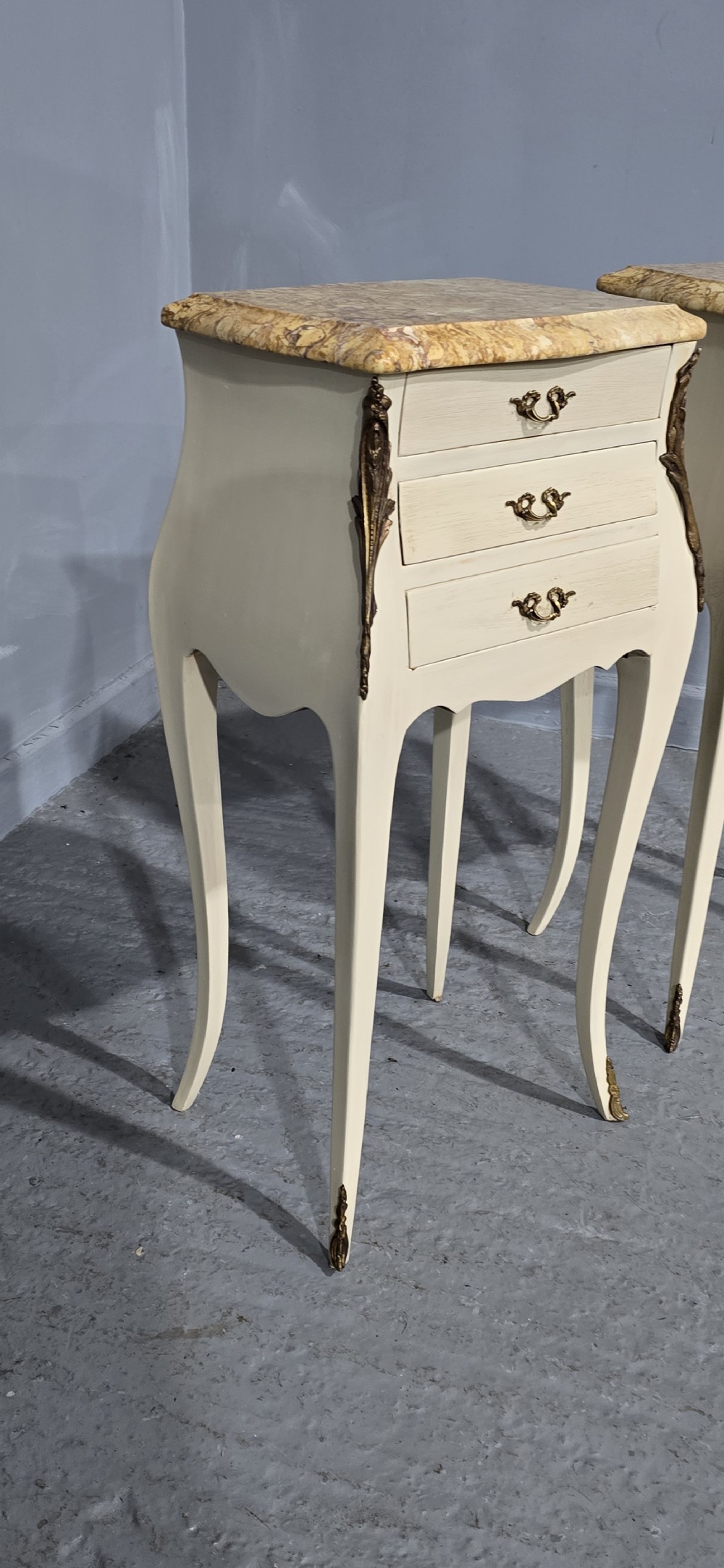 A pair of slender cream-painted French Louis style bedside chests featuring shaped marble tops, three drawers, and decorative brass mounts on tall cabriole legs. View V
