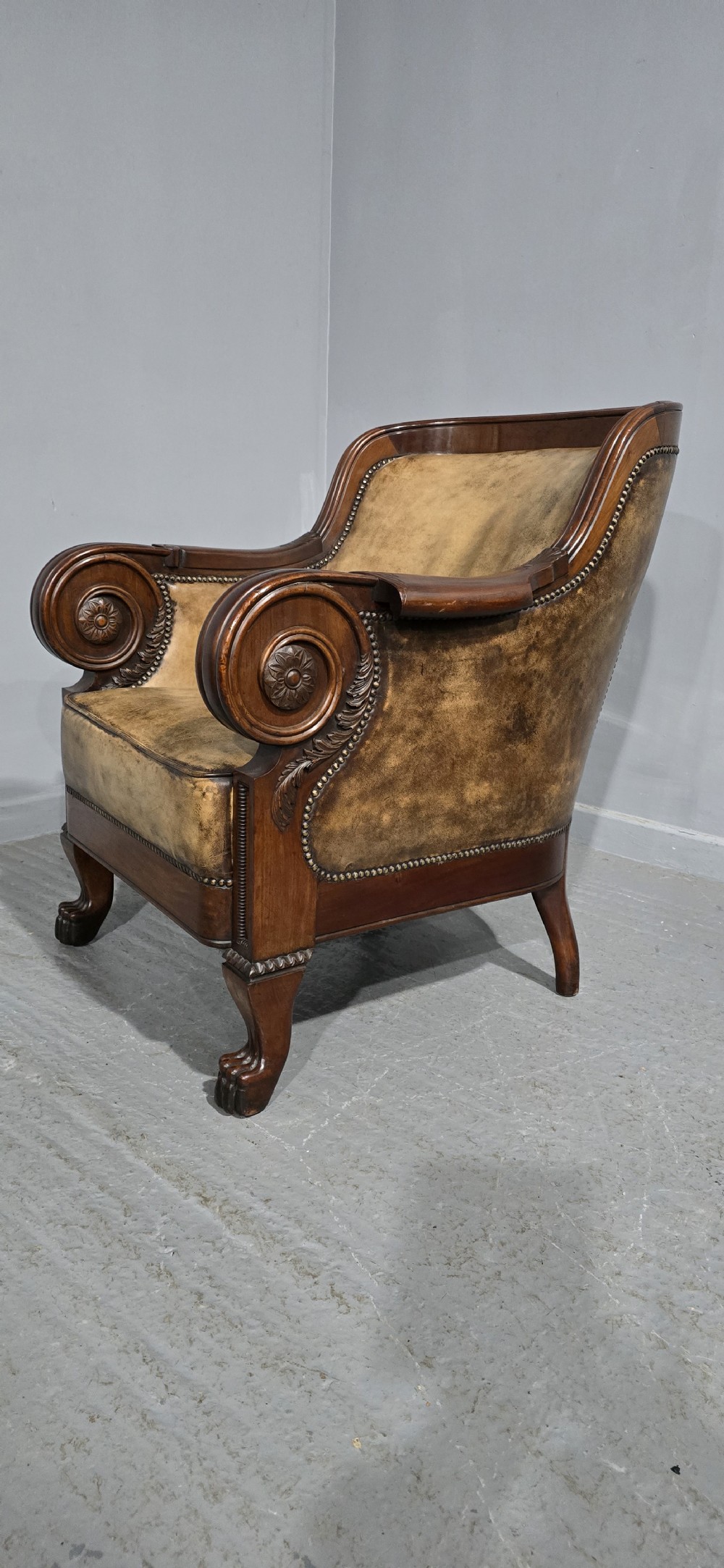 A Regency mahogany library armchair upholstered in aged tan leather featuring carved scroll arms, brass studding, and carved paw feet. View IV
