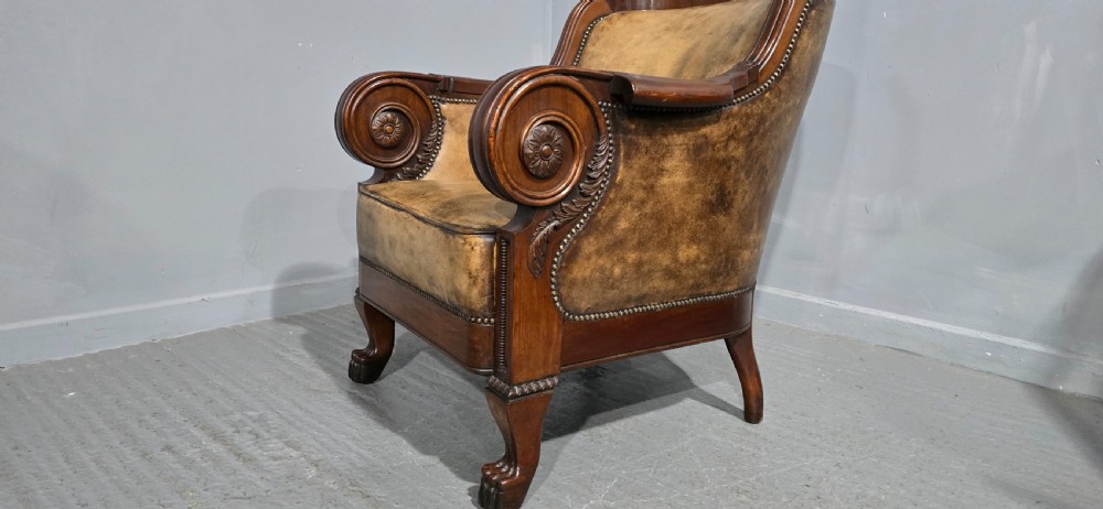 A Regency mahogany library armchair upholstered in aged tan leather featuring carved scroll arms, brass studding, and carved paw feet. View VII