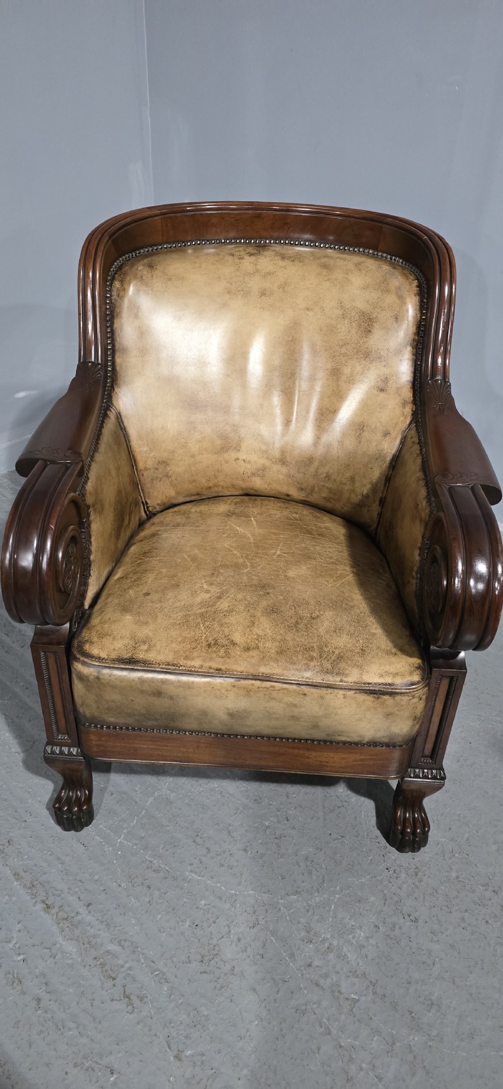 A Regency mahogany library armchair upholstered in aged tan leather featuring carved scroll arms, brass studding, and carved paw feet. View X