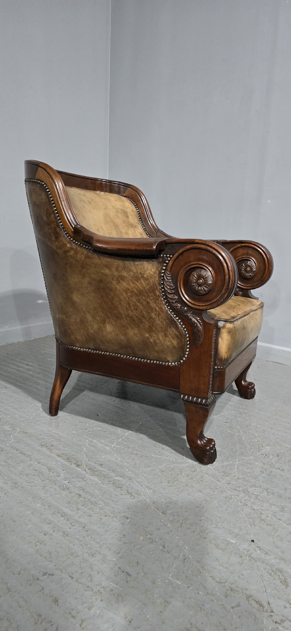 A Regency mahogany library armchair upholstered in aged tan leather featuring carved scroll arms, brass studding, and carved paw feet. View XII