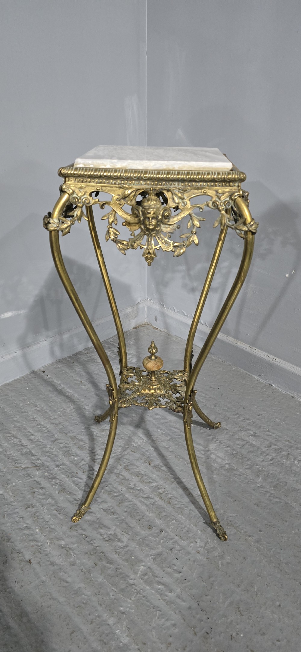 An ornate tall brass plant stand featuring a square white marble top, detailed mask motifs, curved legs, and a lower decorative tier. View II