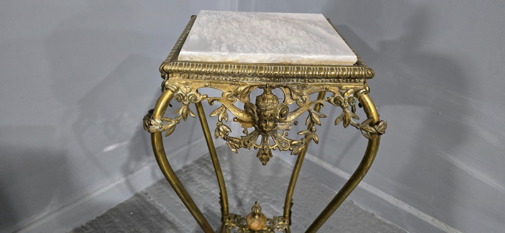 An ornate tall brass plant stand featuring a square white marble top, detailed mask motifs, curved legs, and a lower decorative tier. View III
