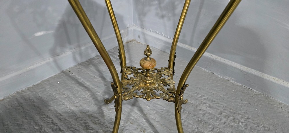 An ornate tall brass plant stand featuring a square white marble top, detailed mask motifs, curved legs, and a lower decorative tier. View V