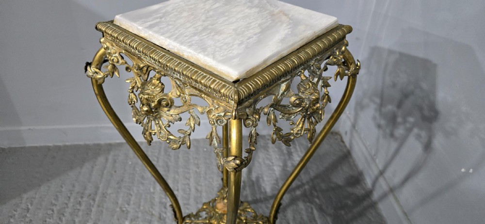 An ornate tall brass plant stand featuring a square white marble top, detailed mask motifs, curved legs, and a lower decorative tier. View VII