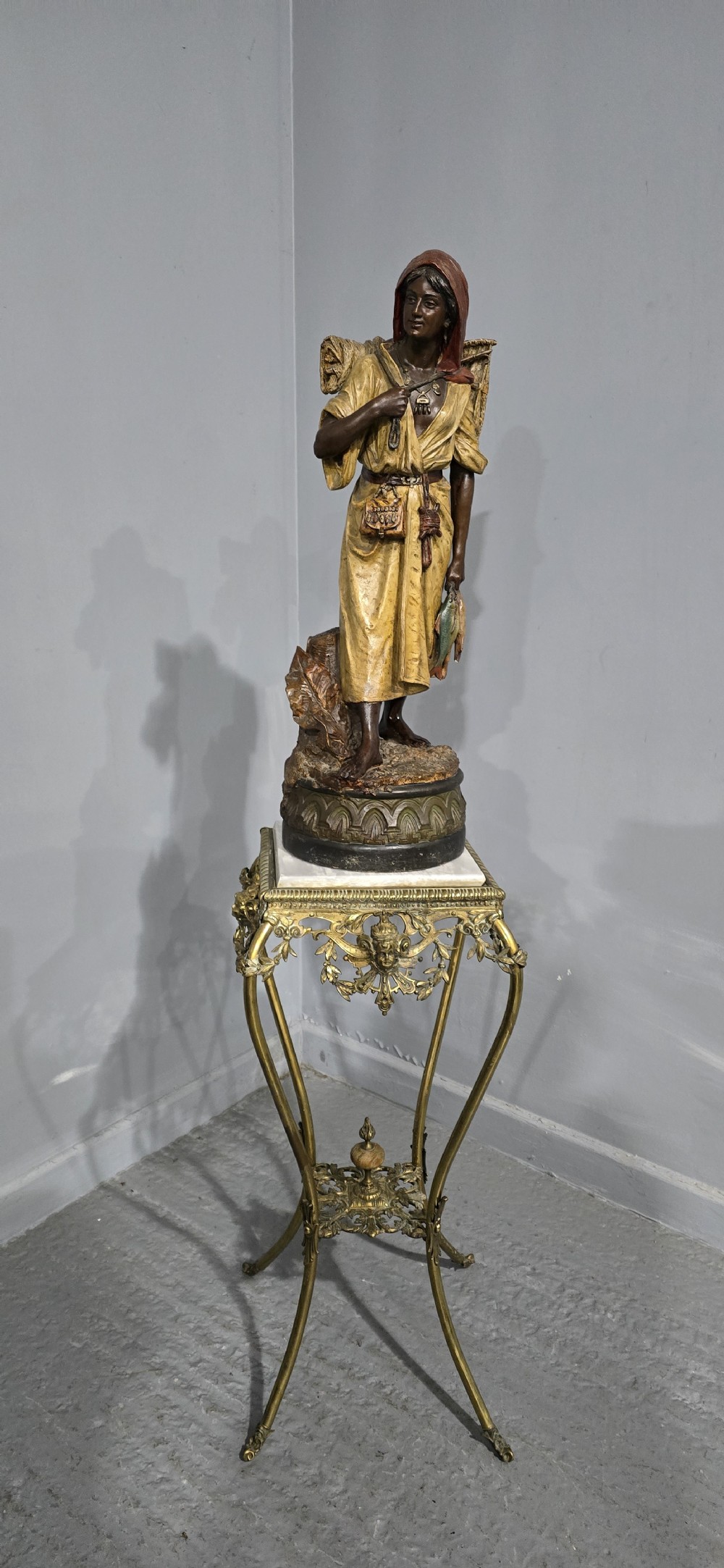 A polychrome terracotta sculpture depicting a standing woman dressed as a fisher girl carrying a basket on her back and holding fish.