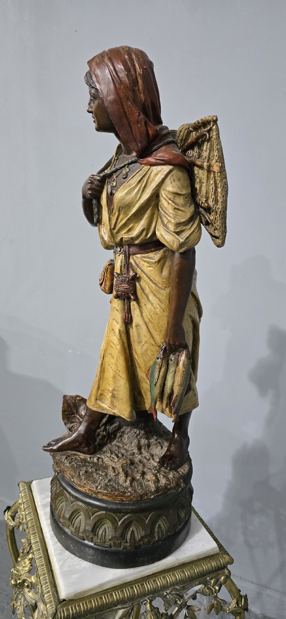 A polychrome terracotta sculpture depicting a standing woman dressed as a fisher girl carrying a basket on her back and holding fish. View III