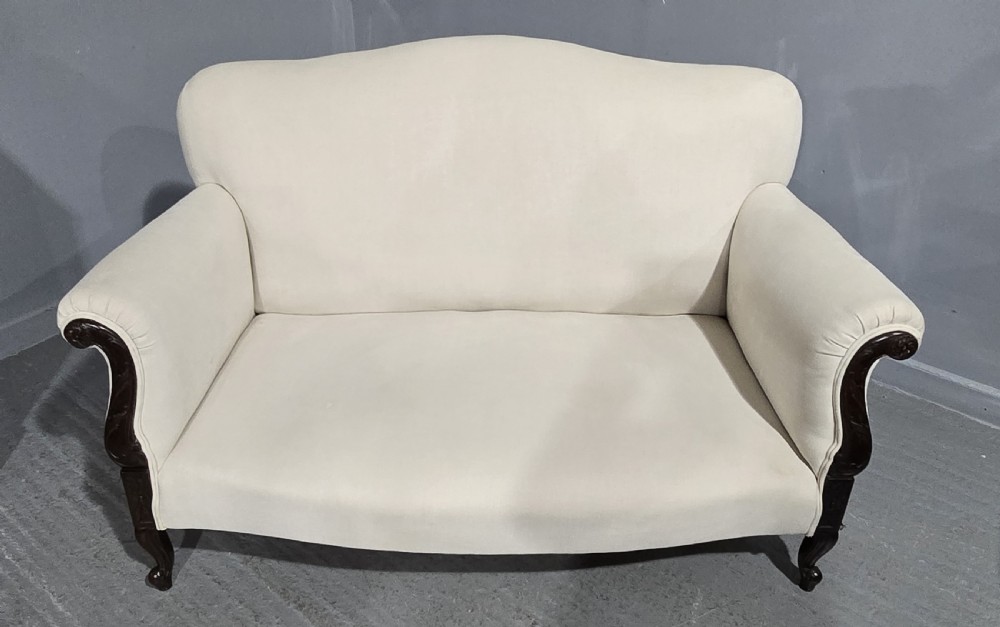 A Victorian two-seater sofa upholstered in plain cream fabric featuring an arched back, rolled padded arms, and dark carved mahogany front legs.
