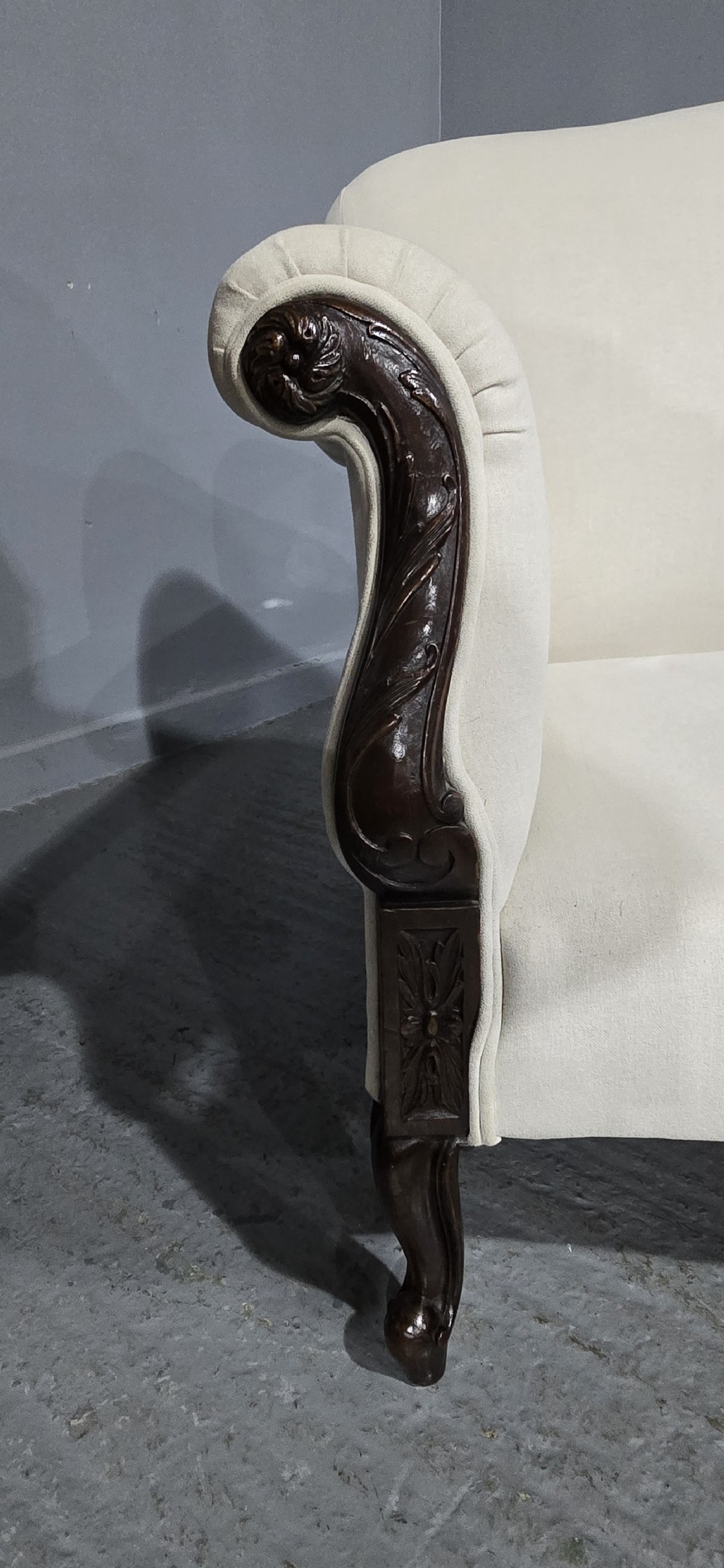A Victorian two-seater sofa upholstered in plain cream fabric featuring an arched back, rolled padded arms, and dark carved mahogany front legs. View V