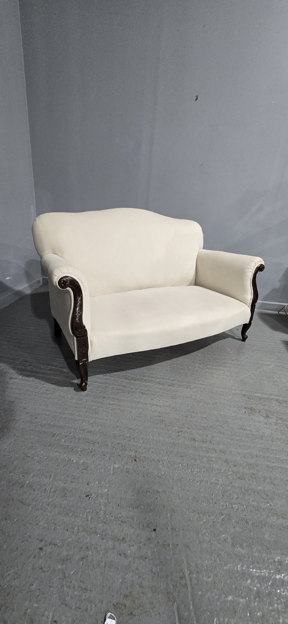 A Victorian two-seater sofa upholstered in plain cream fabric featuring an arched back, rolled padded arms, and dark carved mahogany front legs. View VI