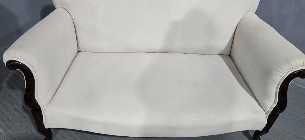 A Victorian two-seater sofa upholstered in plain cream fabric featuring an arched back, rolled padded arms, and dark carved mahogany front legs. View VII