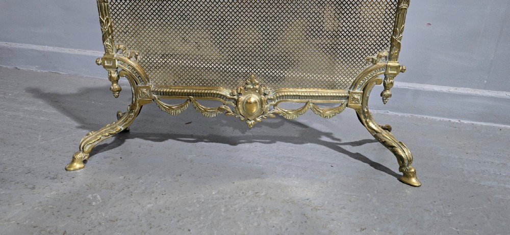 A Victorian gilt brass fire guard featuring a fine mesh panel, ribbon garlands, top wreath crest, and ornate torch finials. View II