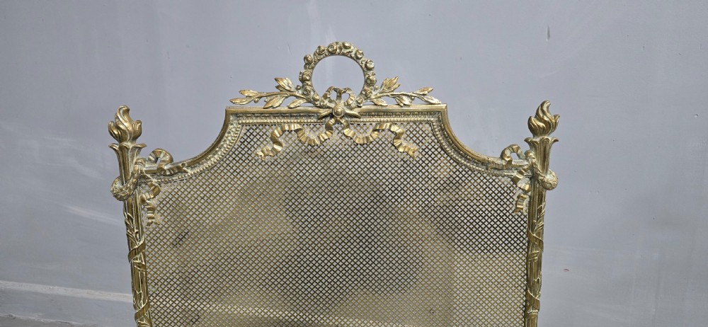 A Victorian gilt brass fire guard featuring a fine mesh panel, ribbon garlands, top wreath crest, and ornate torch finials. View IV