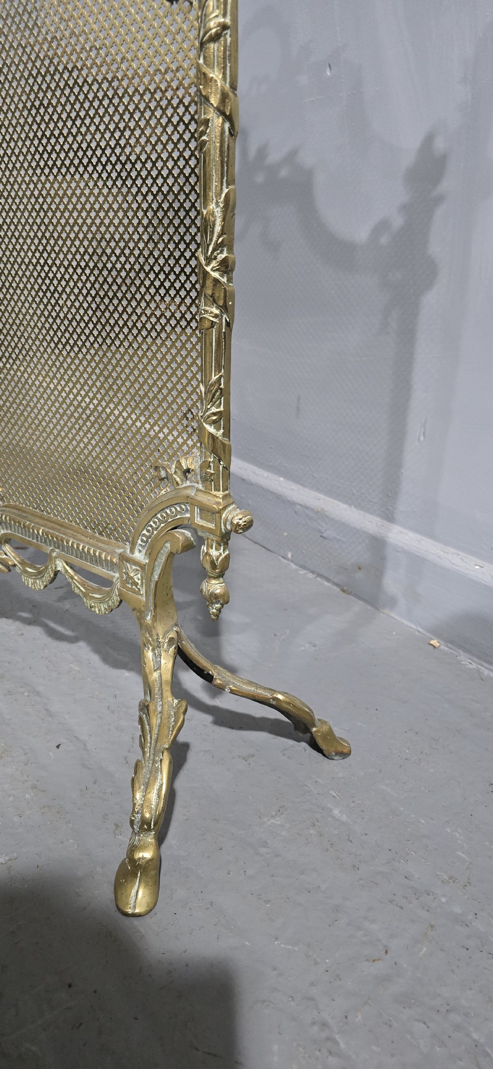 A Victorian gilt brass fire guard featuring a fine mesh panel, ribbon garlands, top wreath crest, and ornate torch finials. View V