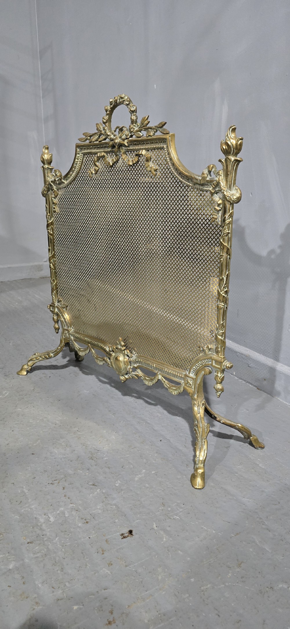 A Victorian gilt brass fire guard featuring a fine mesh panel, ribbon garlands, top wreath crest, and ornate torch finials. View VI