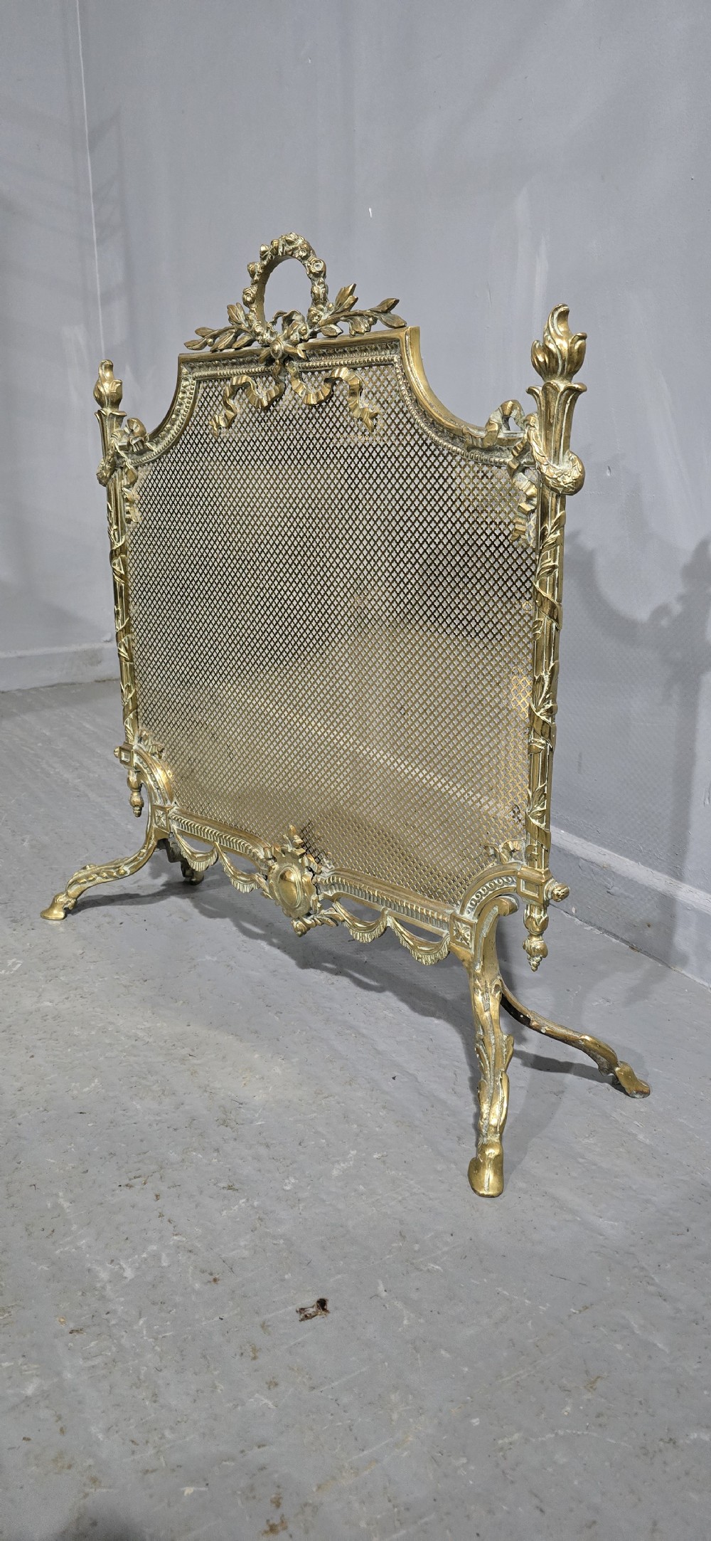 A Victorian gilt brass fire guard featuring a fine mesh panel, ribbon garlands, top wreath crest, and ornate torch finials. View VII