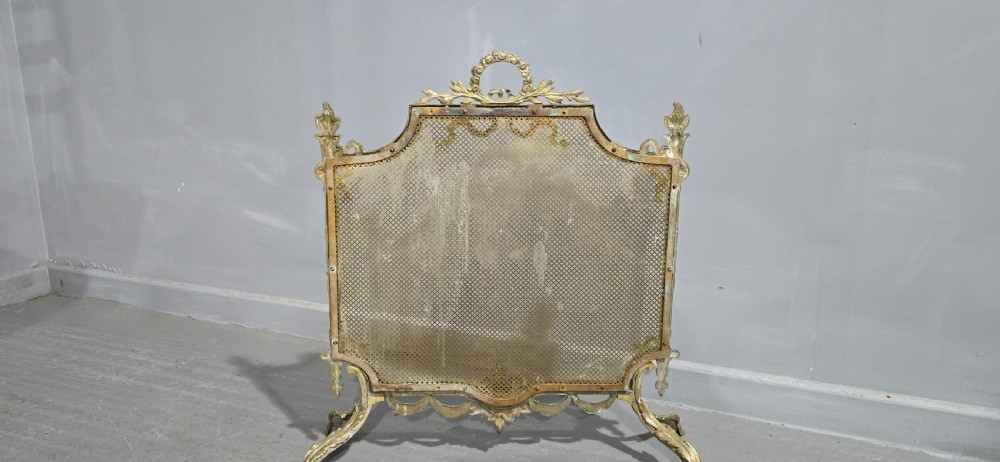A Victorian gilt brass fire guard featuring a fine mesh panel, ribbon garlands, top wreath crest, and ornate torch finials. View VIII