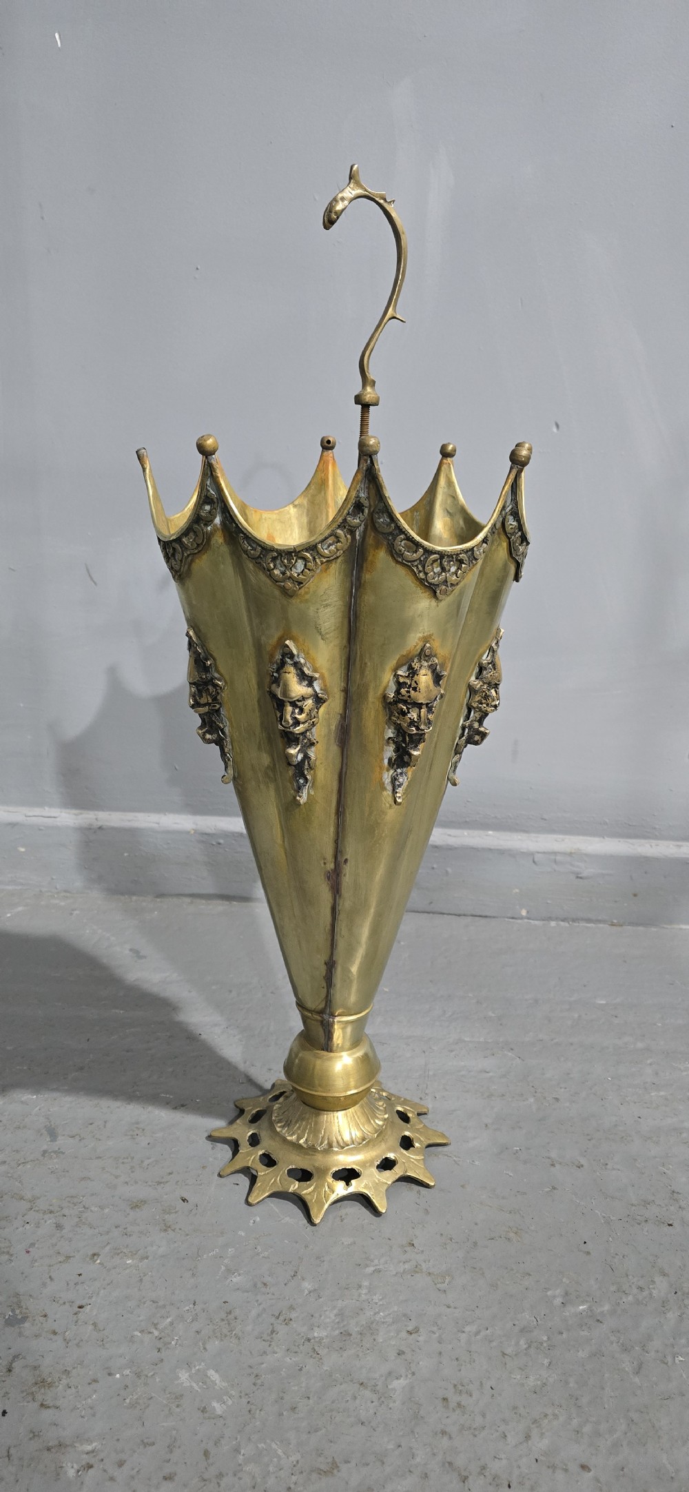 An antique umbrella-shaped brass umbrella stand featuring relief mascaron face details and a central curved hook handle. View II