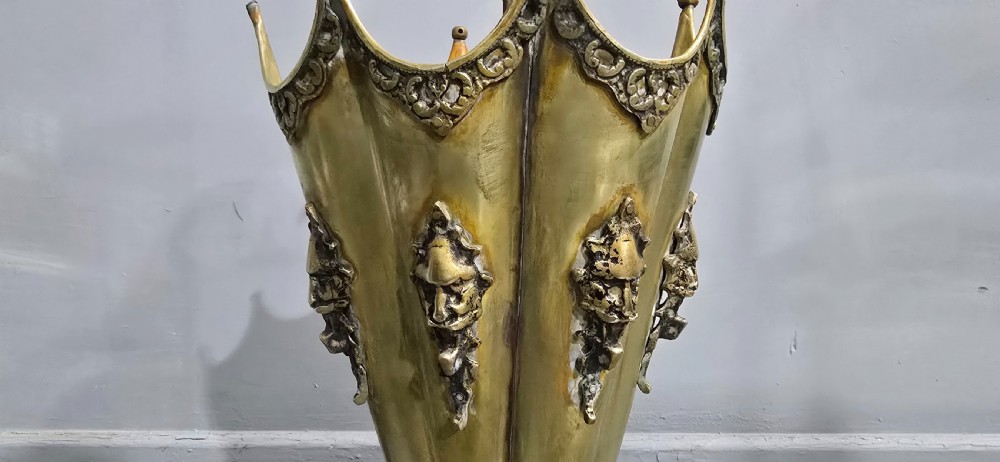 An antique umbrella-shaped brass umbrella stand featuring relief mascaron face details and a central curved hook handle. View III