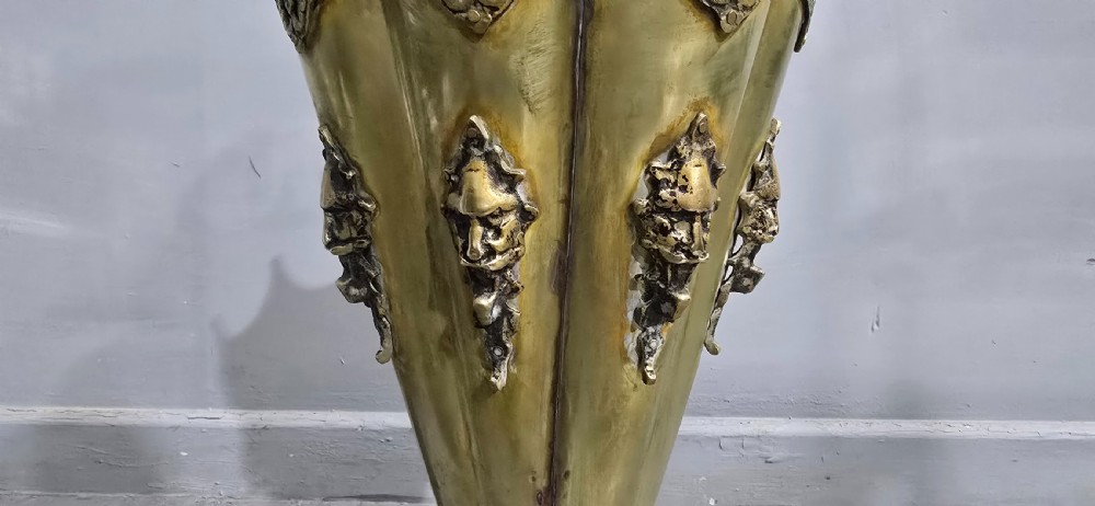An antique umbrella-shaped brass umbrella stand featuring relief mascaron face details and a central curved hook handle. View V