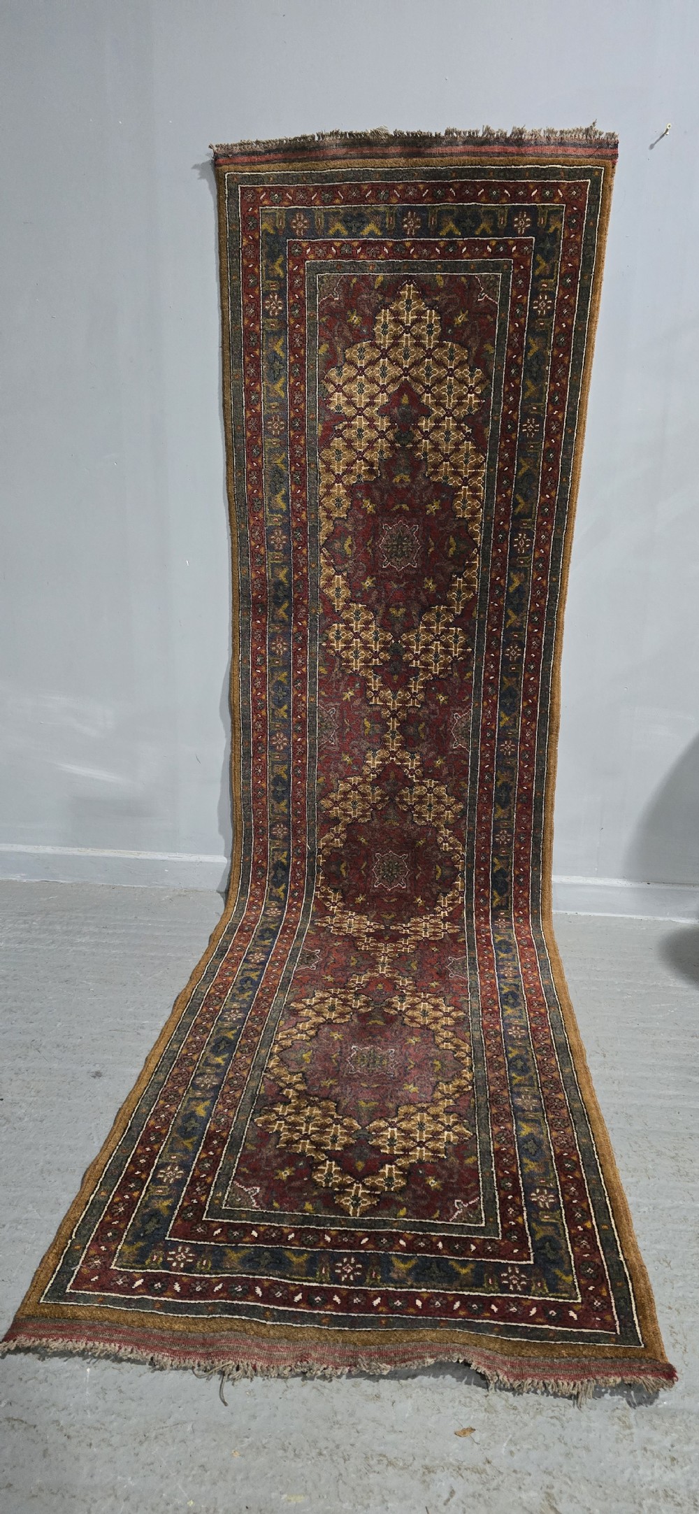 A long, narrow hand-knotted wool runner rug featuring repeating geometric medallions in deep red, navy blue, and ochre with intricate borders and fringed ends.