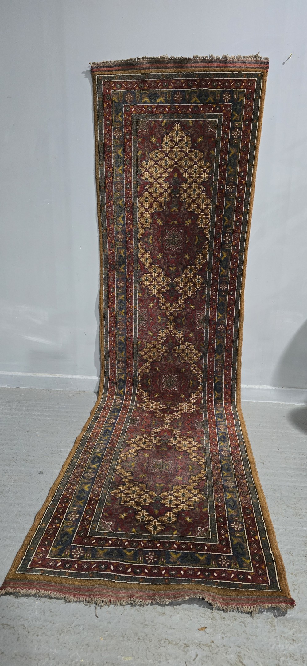 A long, narrow hand-knotted wool runner rug featuring repeating geometric medallions in deep red, navy blue, and ochre with intricate borders and fringed ends. View II