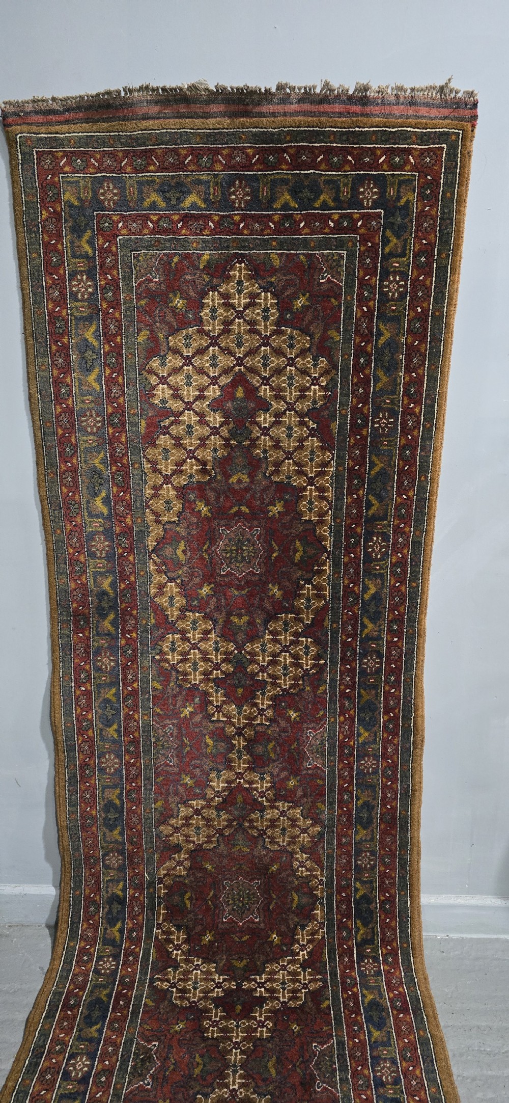 A long, narrow hand-knotted wool runner rug featuring repeating geometric medallions in deep red, navy blue, and ochre with intricate borders and fringed ends. View III