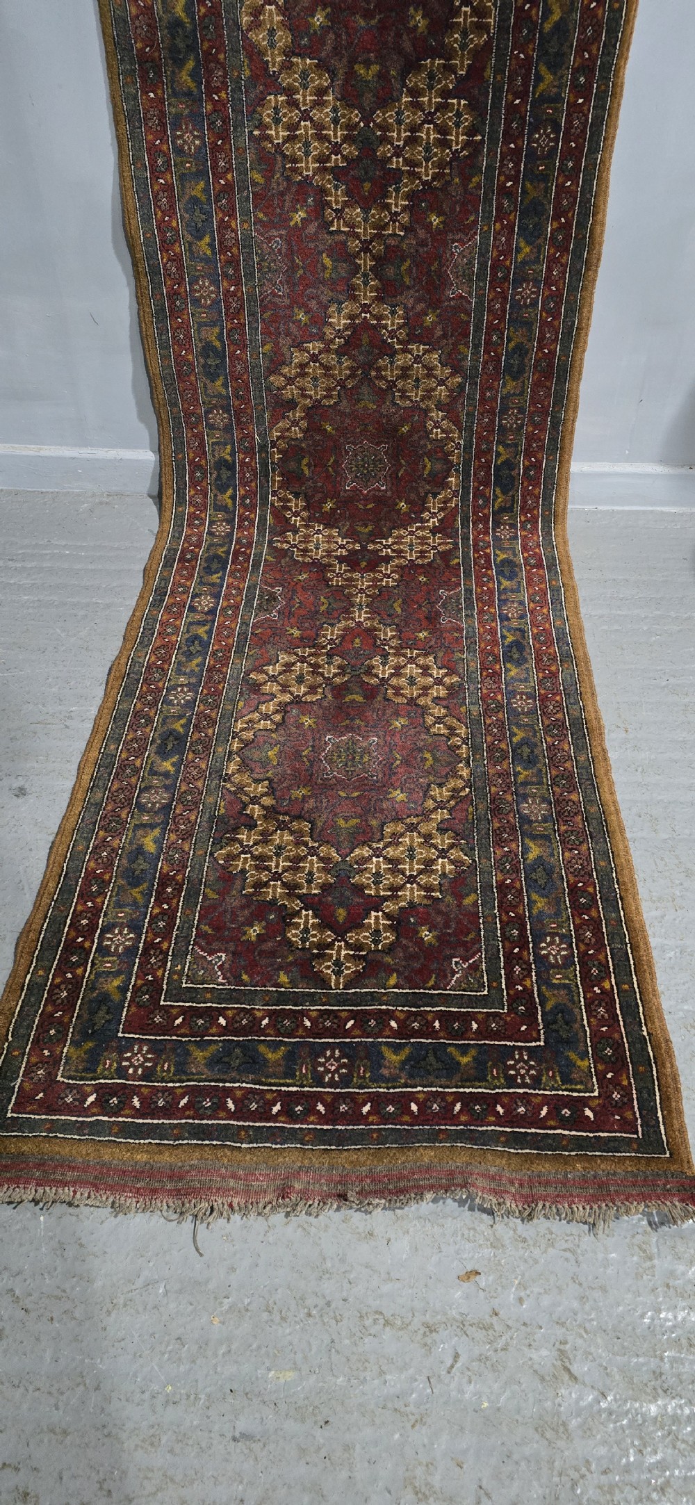 A long, narrow hand-knotted wool runner rug featuring repeating geometric medallions in deep red, navy blue, and ochre with intricate borders and fringed ends. View IV