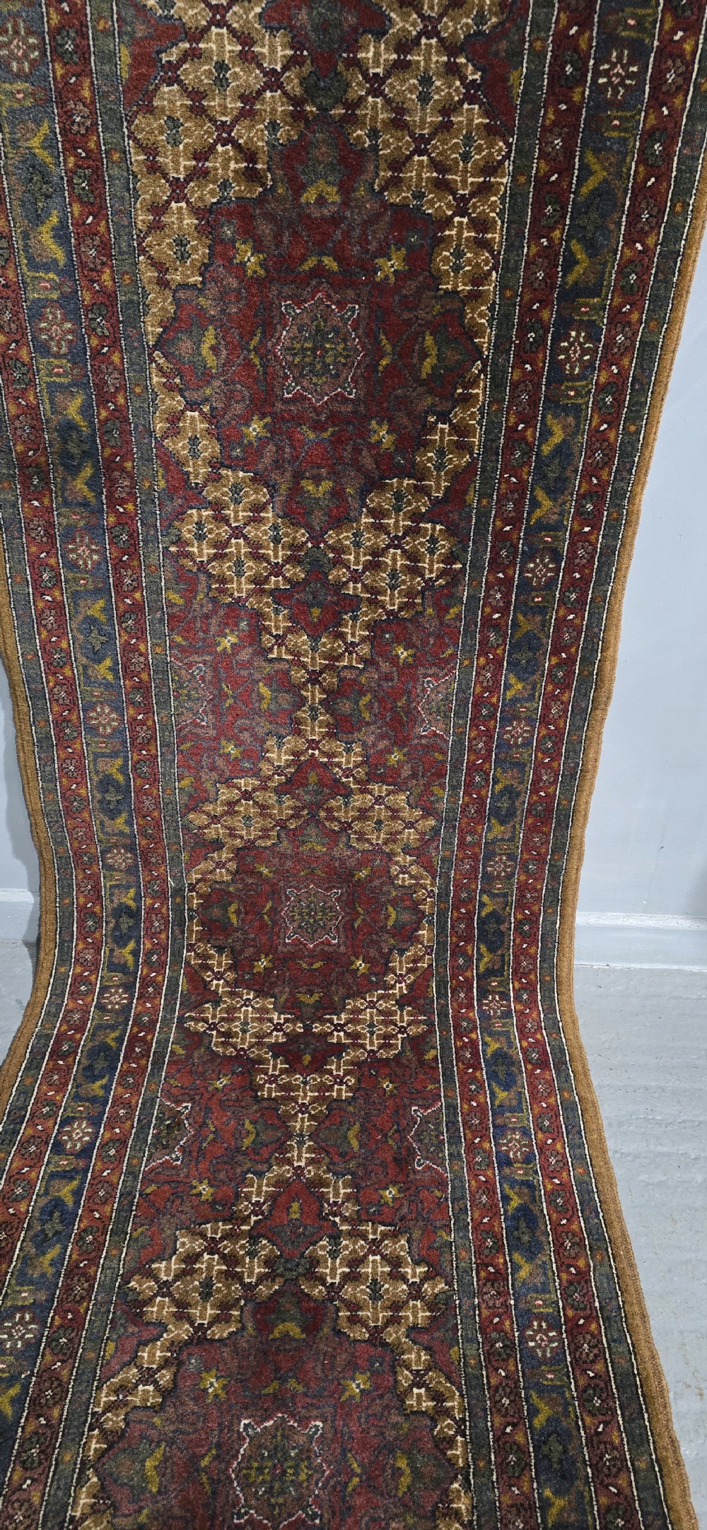 A long, narrow hand-knotted wool runner rug featuring repeating geometric medallions in deep red, navy blue, and ochre with intricate borders and fringed ends. View V