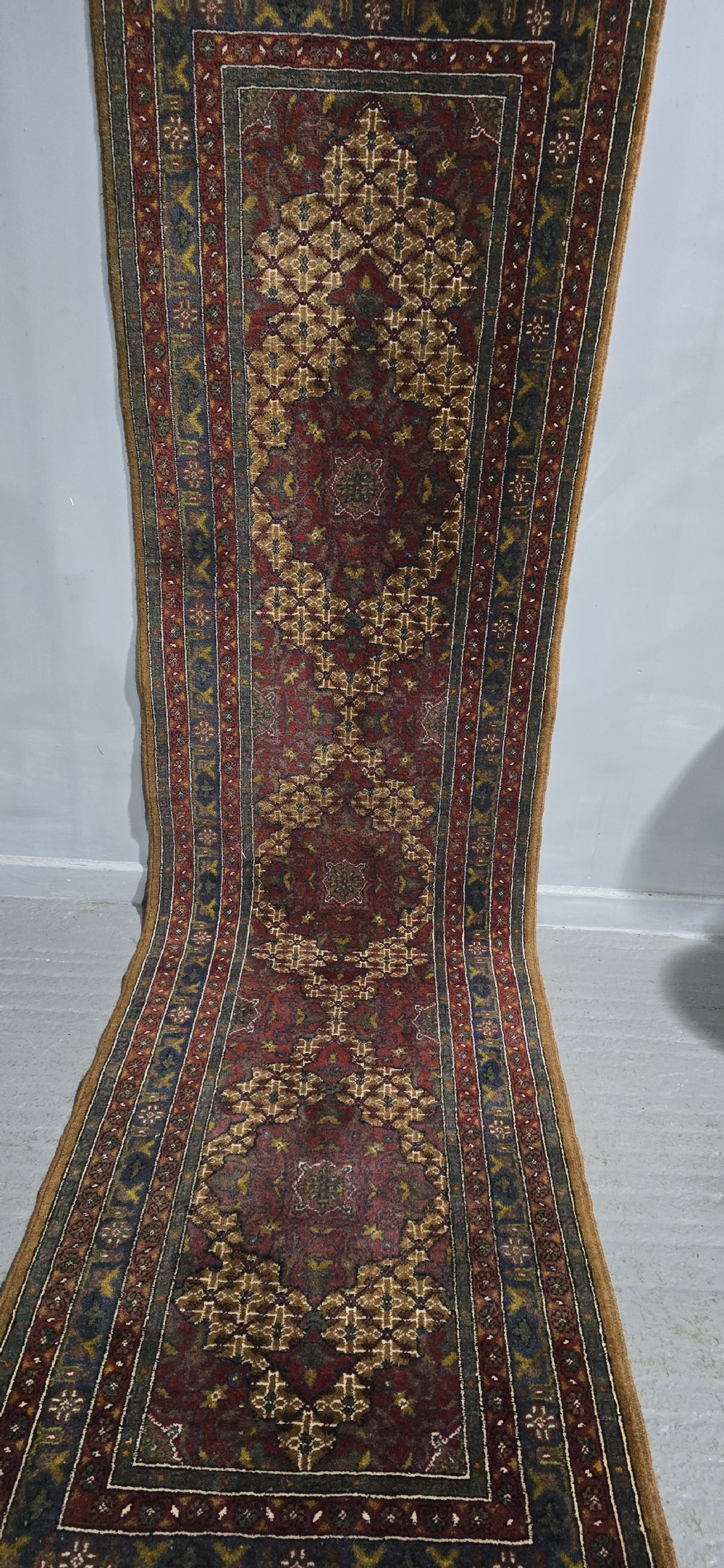 A long, narrow hand-knotted wool runner rug featuring repeating geometric medallions in deep red, navy blue, and ochre with intricate borders and fringed ends. View VI