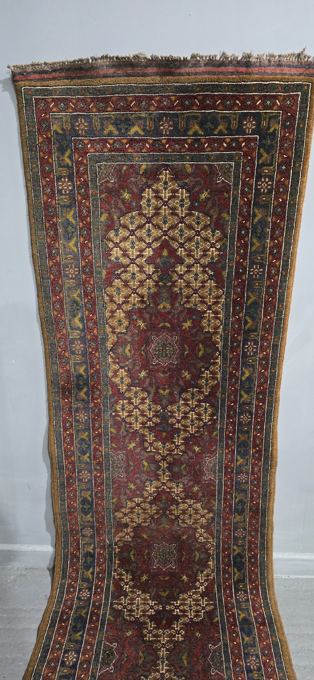 A long, narrow hand-knotted wool runner rug featuring repeating geometric medallions in deep red, navy blue, and ochre with intricate borders and fringed ends. View VIII