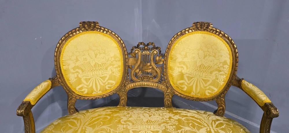 An antique French Louis XVI style giltwood two-seat sofa with twin oval medallion backrests, a central carved lyre motif, fluted legs, and yellow silk damask upholstery. View V
