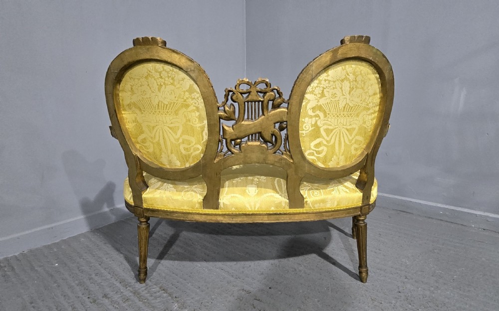 An antique French Louis XVI style giltwood two-seat sofa with twin oval medallion backrests, a central carved lyre motif, fluted legs, and yellow silk damask upholstery. View XIV