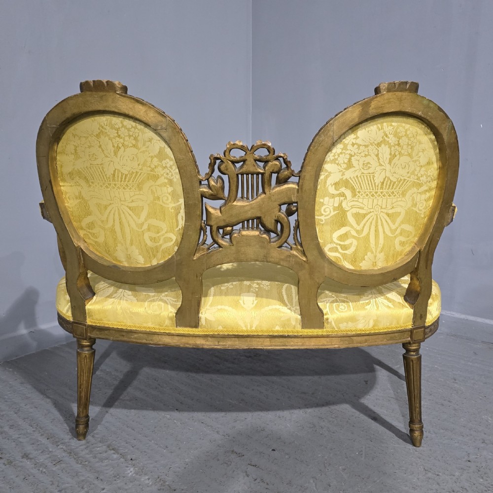 An antique French Louis XVI style giltwood two-seat sofa with twin oval medallion backrests, a central carved lyre motif, fluted legs, and yellow silk damask upholstery. View XV