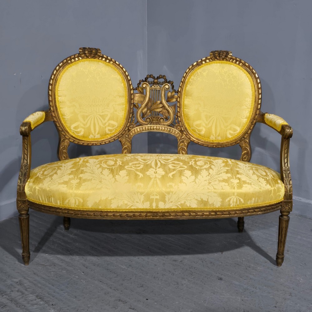 An antique French Louis XVI style giltwood two-seat sofa with twin oval medallion backrests, a central carved lyre motif, fluted legs, and yellow silk damask upholstery.