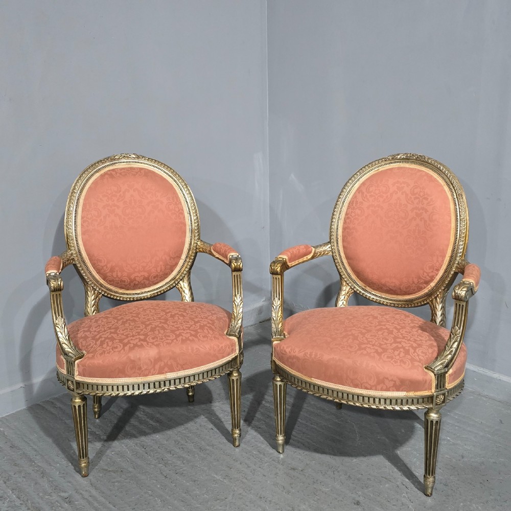 A pair of French Louis XVI style elbow chairs featuring silver gilt carved frames with oval backs, fluted legs, and pink patterned upholstery.