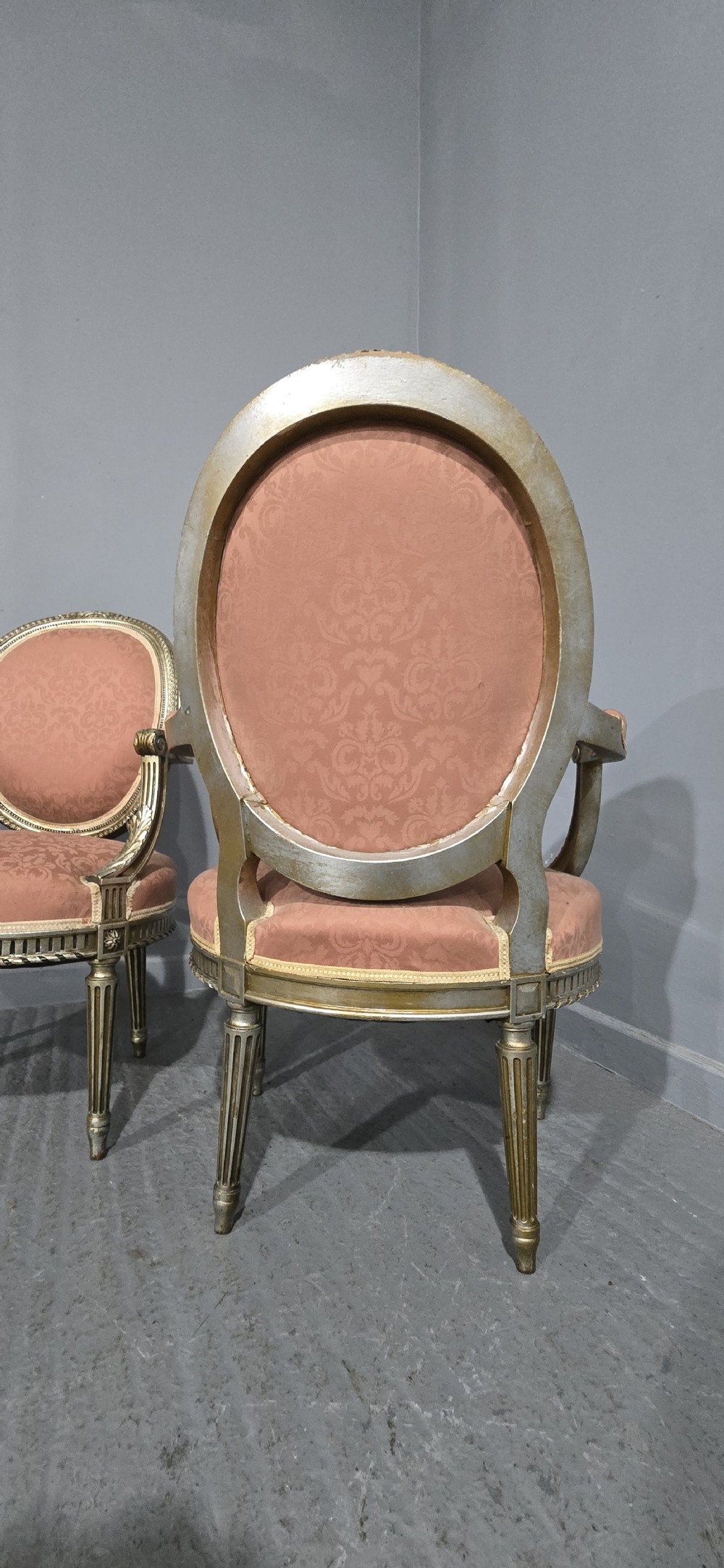 A pair of French Louis XVI style elbow chairs featuring silver gilt carved frames with oval backs, fluted legs, and pink patterned upholstery. View X