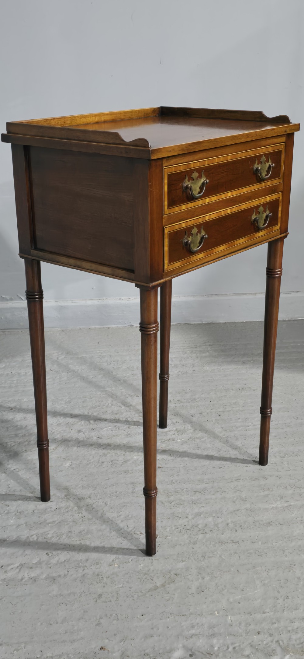 A dark mahogany side table featuring two drawers with brass batwing drop handles, crossbanded inlay borders, and slender turned legs. View VII