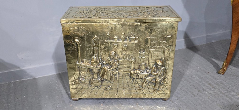 A rectangular Victorian brass log bin featuring detailed repousse relief panels of figures inside a domestic hearth interior. View III