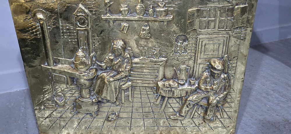 A rectangular Victorian brass log bin featuring detailed repousse relief panels of figures inside a domestic hearth interior. View IV