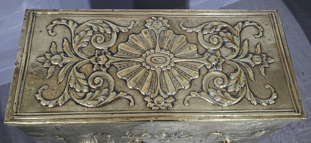 A rectangular Victorian brass log bin featuring detailed repousse relief panels of figures inside a domestic hearth interior. View V
